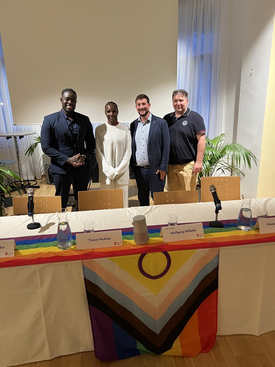 Panelists seated behind a table with a progress Pride flag